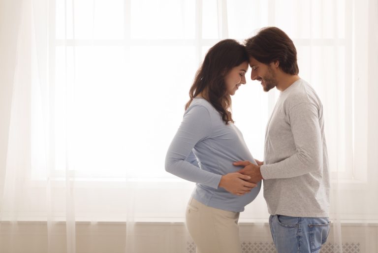 young couple expecting baby standing together against window at home