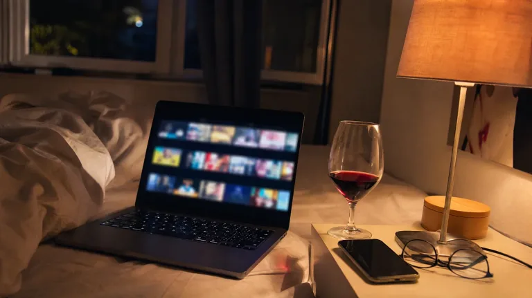 Laptop auf zerwühltem Bett mit verschwommenen Video-Vorschaubildern, daneben Rotweinglas, Smartphone und Brille auf dem Nachttisch im Lampenlicht