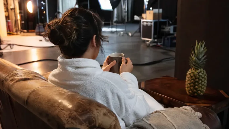 Darstellerin im Bademantel macht mit einer Tasse Tee eine Drehpause auf einem Ledersofa im Filmstudio, daneben eine Ananas